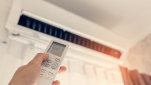 Person's hand aiming a remote at a ductless mini-split installed high on a white wall, above a window.