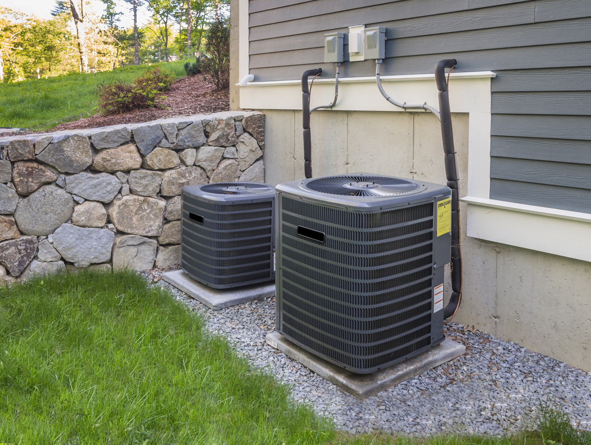 Two residential HVAC units near a gray suburban house with stone retaining wall and mowed green yard in background.