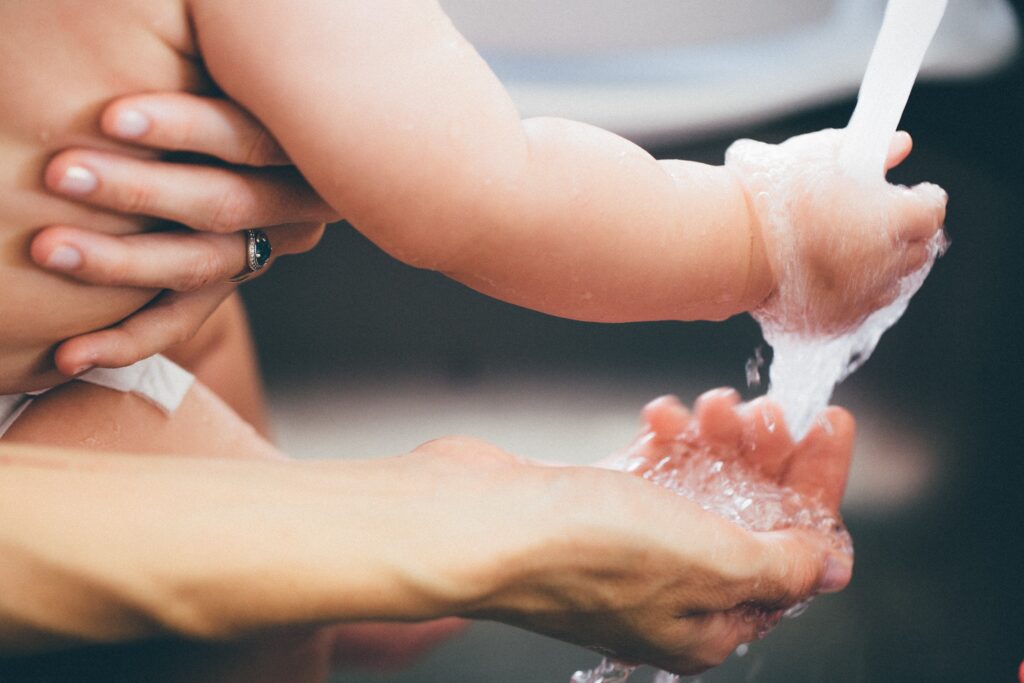 Close view of a woman holding a baby; both with their hands under a running faucet.