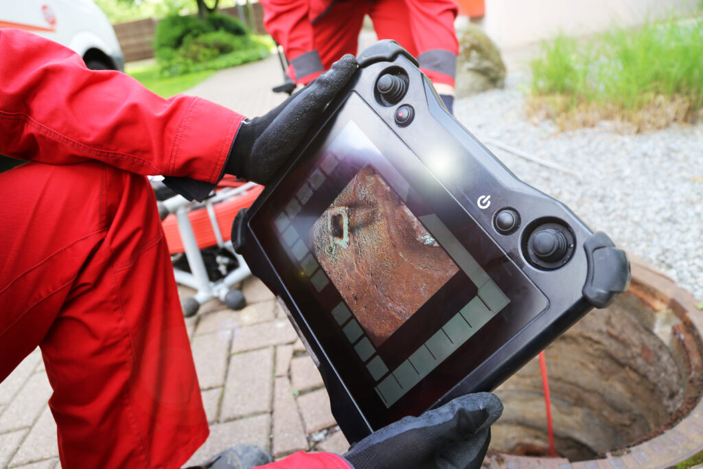Close view of a plumber's hand holding a sewer line inspection camera, showing view of the interior of a sewer pipe.