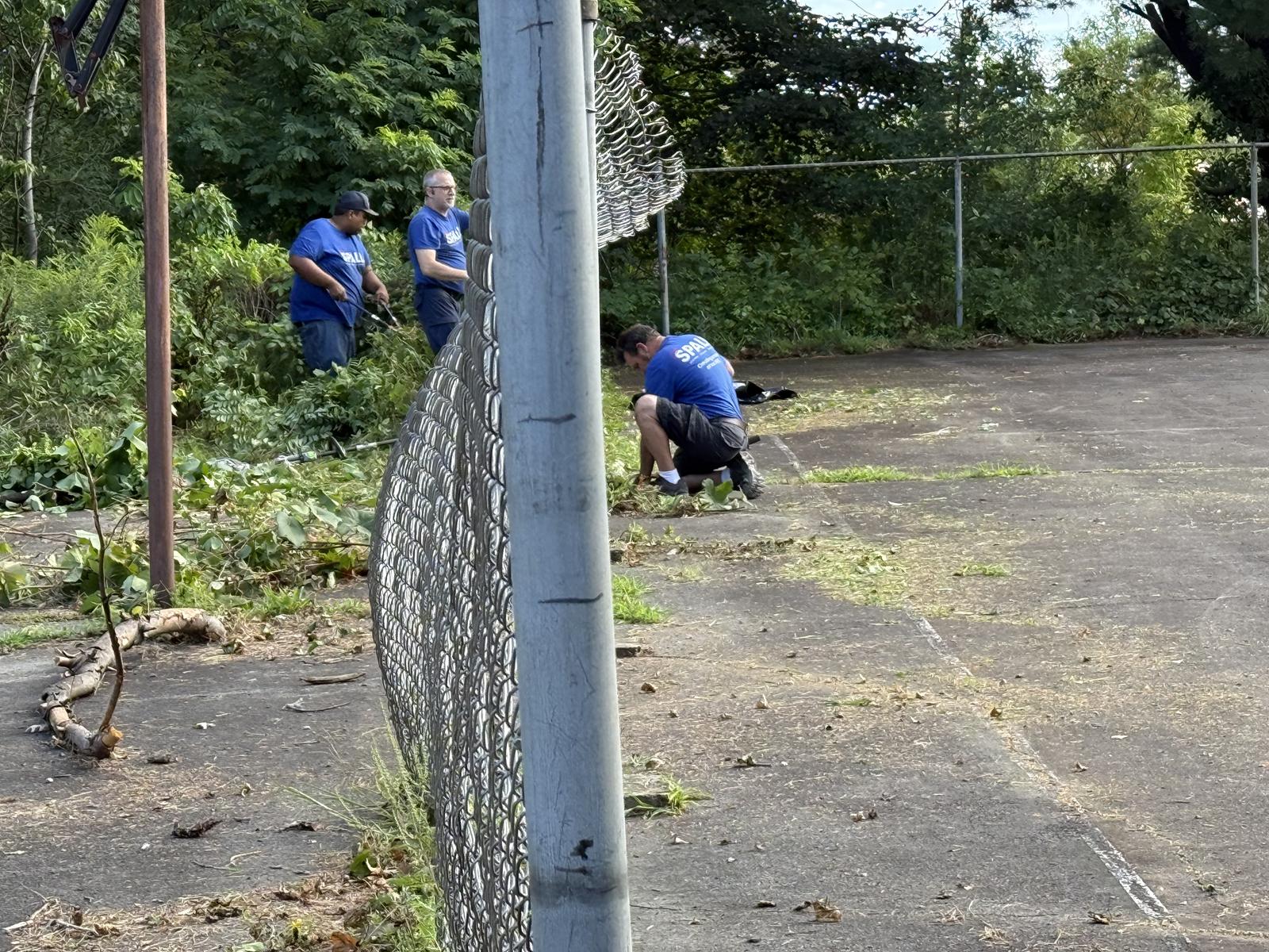 Spall crew members cutting back overgrowth along the park fence line.